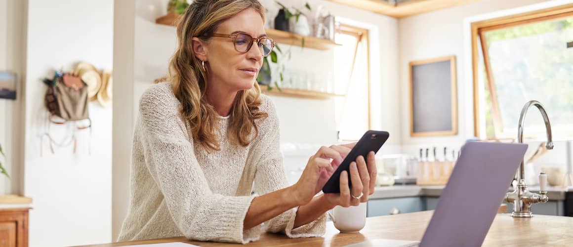 Blonde woman wearing glasses using her phone in the kitchen in front of a laptop.