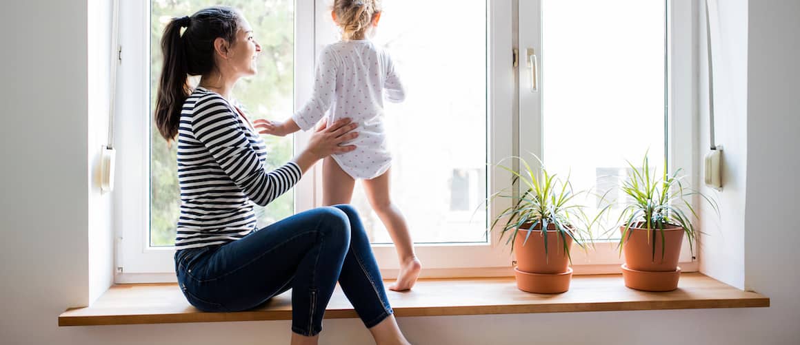 Image of mom helping young child look out the window, while standing on window seat.