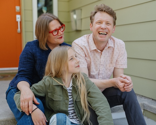 Family sitting on front porch steps of green home with orange door.