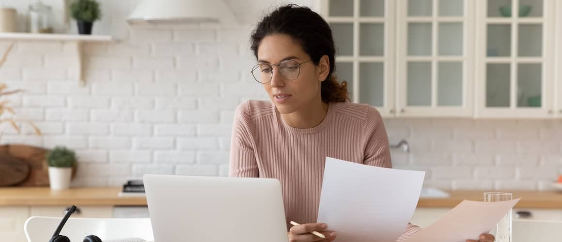 Young woman with a neutral expression, holding papers in hand while looking at a laptop in the kitchen.