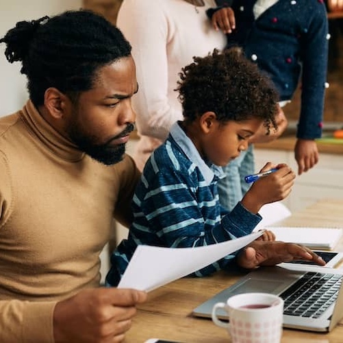 Father at kitchen table with young son on his lap reviewing paperwork and information on laptop..