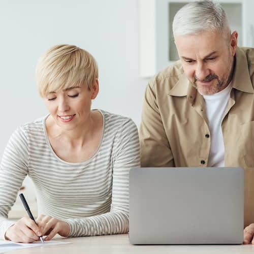 A middle-aged couple at a kitchen island using a computer and calculator, potentially managing finances or planning for a home purchase.