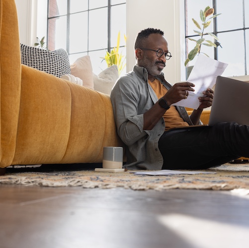 Man lounging on floor of airy living room on laptop looking at mortgage paperwork.