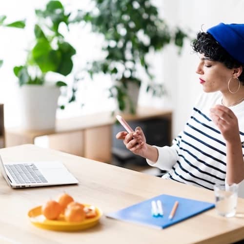 Young woman checking phone at desk with open laptop.