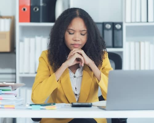 A young woman in a yellow suit coat resting her chin on her hands while deep in thought at her desk.