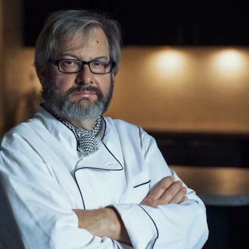 Headshot of a man with grey hair and beard in a chef's coat.