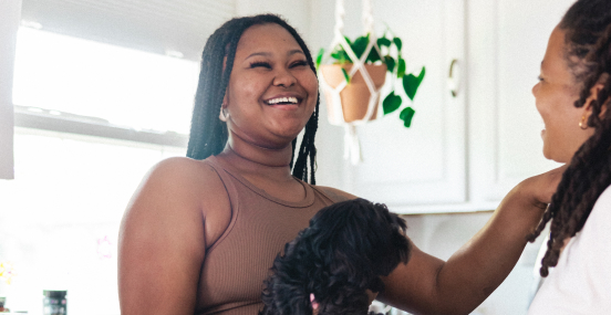 A young Black woman holding a black puppy shares a laugh with a friend in her kitchen.