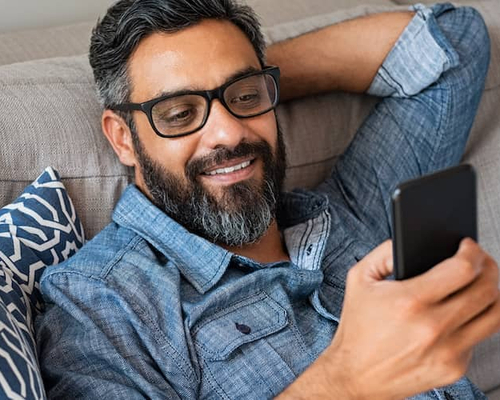 Image of man holding cell phone on couch, smiling at screen.