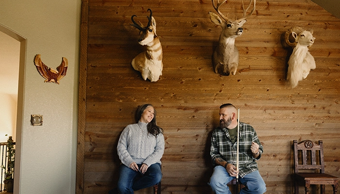 Couple sitting and smiling at each other holding pool sticks under mounted animal heads.