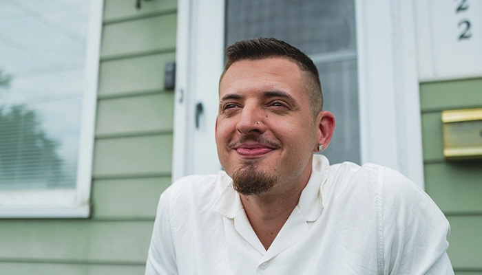 Man in white shirt smiling, sitting in front of sage green house.