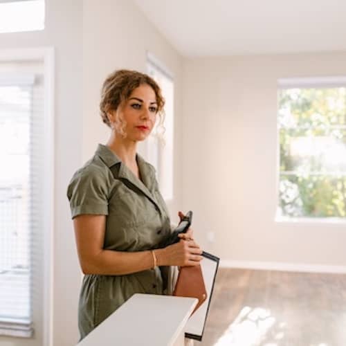 Woman in empty new construction home holding phone and clipboard.