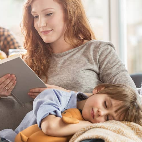 Image of woman reading as sleeping child sits on her lap. Man on phone in background.