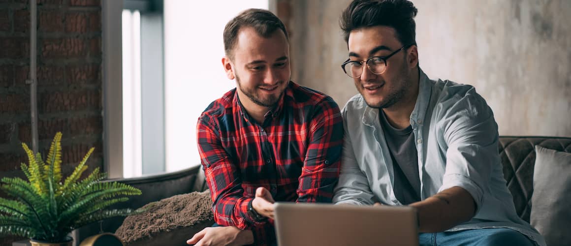 A same-sex male couple spending time together at home with a laptop and coffee, representing inclusivity in homeownership imagery.