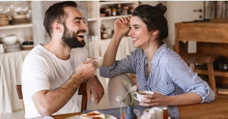 A smiling couple sits at the table in a kitchen drinking coffee.