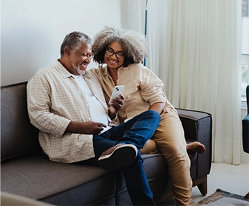 An older African American couple relaxing on a couch and looking at a phone, smiling.