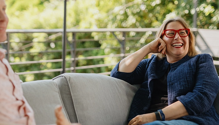 Woman with red glasses resting her feet on another person's lap on outdoor furniture.