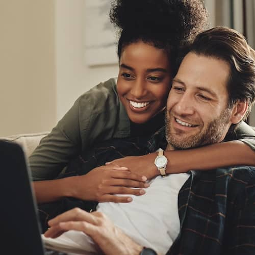 Couple snuggling on couch and reviewing information online on open laptop.