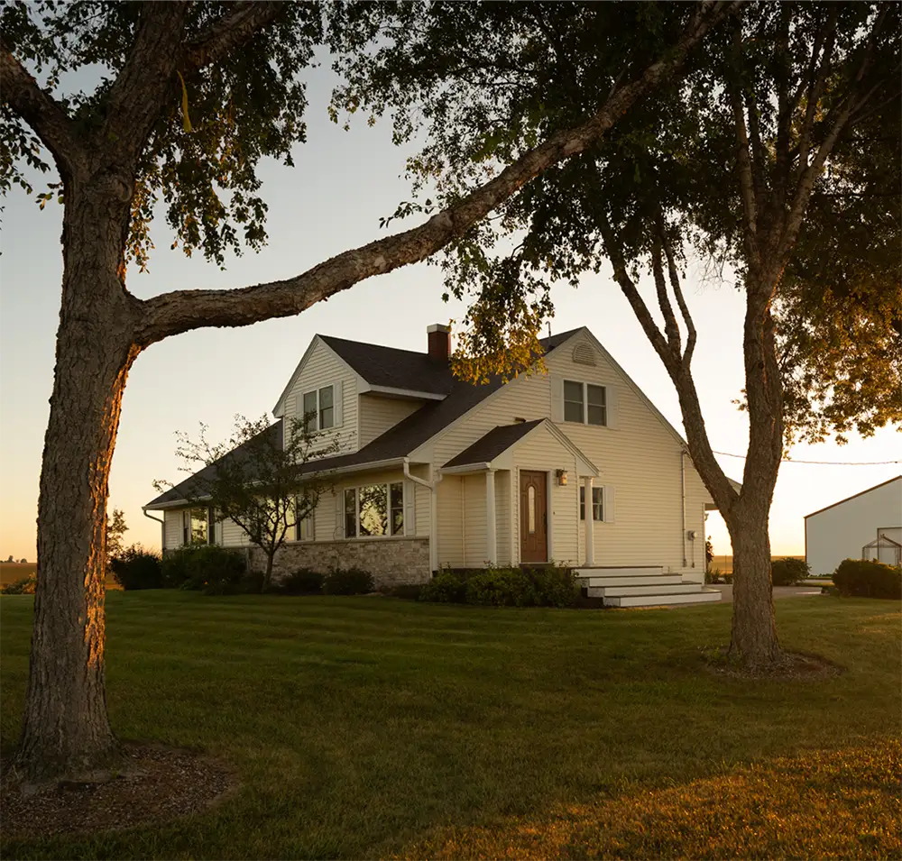 A home on a large field of land, surrounded by trees and a sunset reflecting off the house.