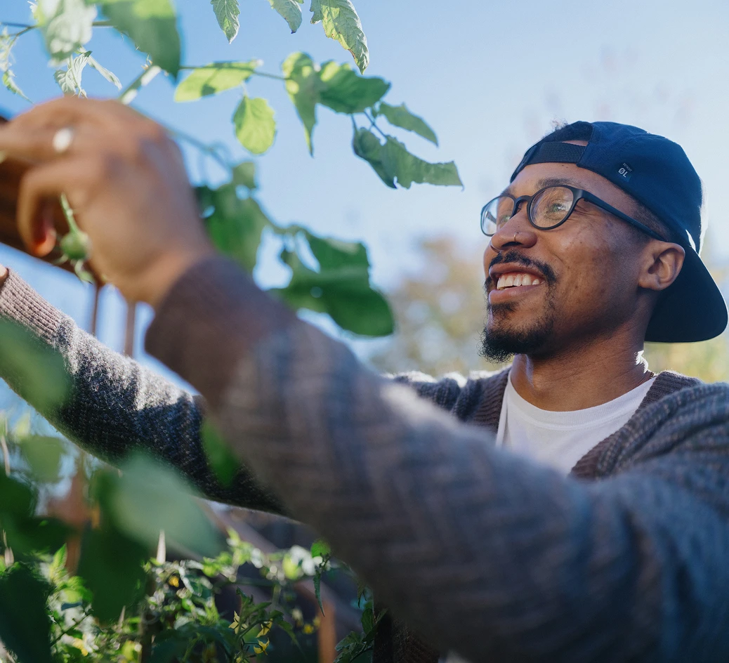 Black man with glasses smiles while reaching up to tend a tall garden plant