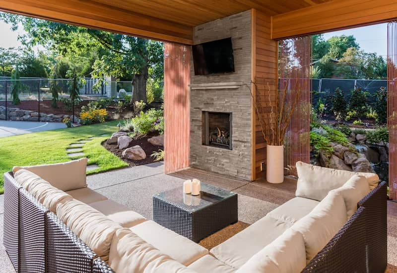 Beautiful covered patio outside new luxury home with television, fireplace, and lush green yard