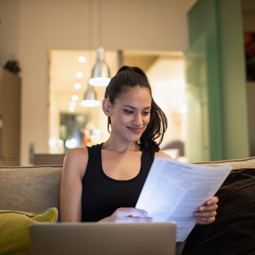 Woman smiling, reviwing paperwork with open laptop in her living room.