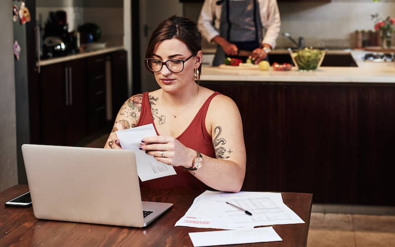 Woman at kitchen table in front of laptop doing bills while partner cooks and chops vegetables in the background.