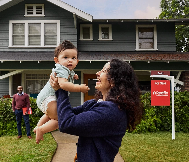 A woman lifting a baby in front of a green two-story house with a Redfin “Sold” sign in the yard while the realtor stands behind them.