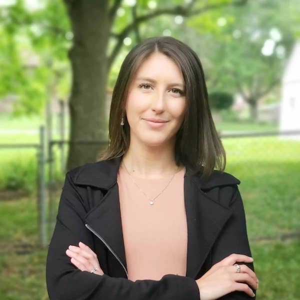 Headshot of Lauren Slagter wearing black leather jacket with arms crossed standing outside on a green lawn by a tree.