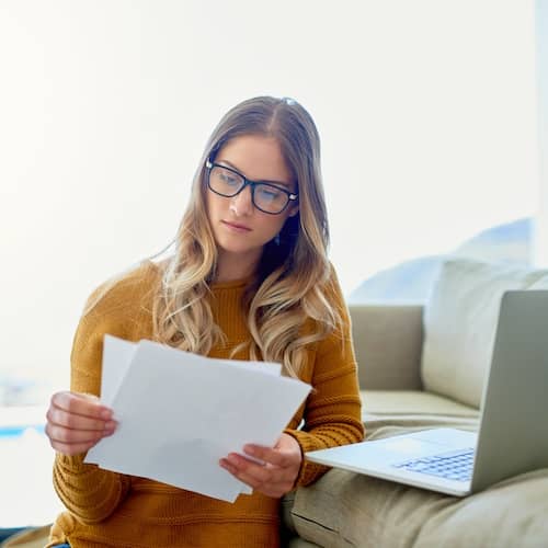 A woman reviews paperwork while sitting on the floor next to her couch where an open laptop rests.