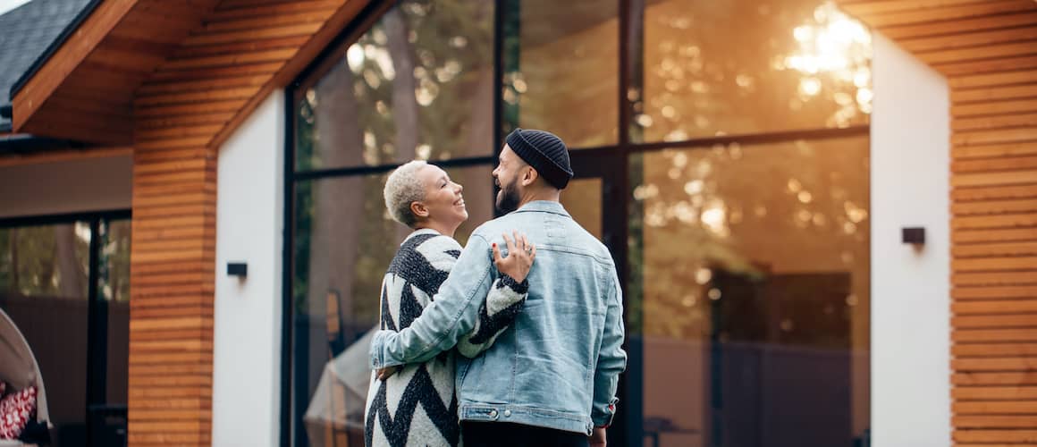 Older African American couple side hugging outside home.