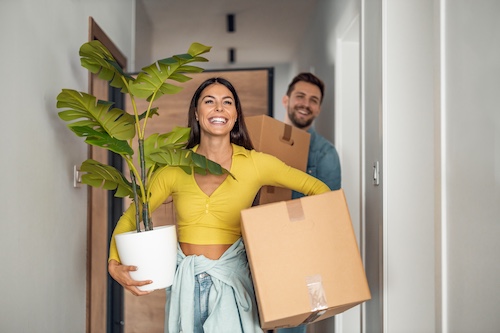 Young couple moving into new home, smiling and carrying boxes and a houseplant.