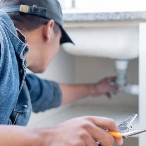 A man inspecting the underside of a kitchen sink while holding a clipboard.