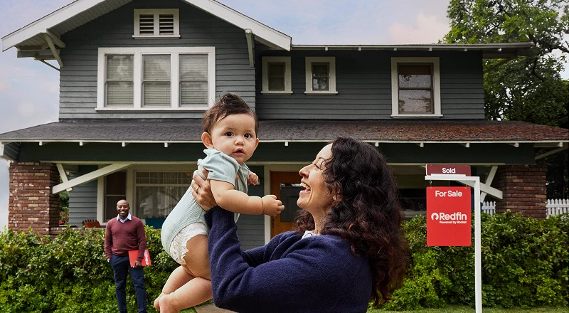 A woman lifting a baby in front of a green two-story house with a Redfin “Sold” sign in the yard while the realtor stands behind them.