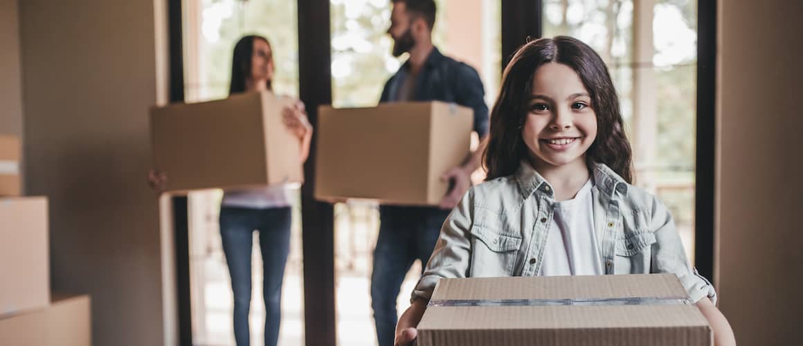 Young girl smiling in foreground, holding a box, moving into new home. Parents are in the background holding boxes and looking at one another, smiling.