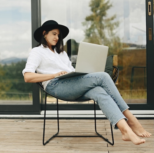 Young woman in stylish hat on laptop on a screen porch,