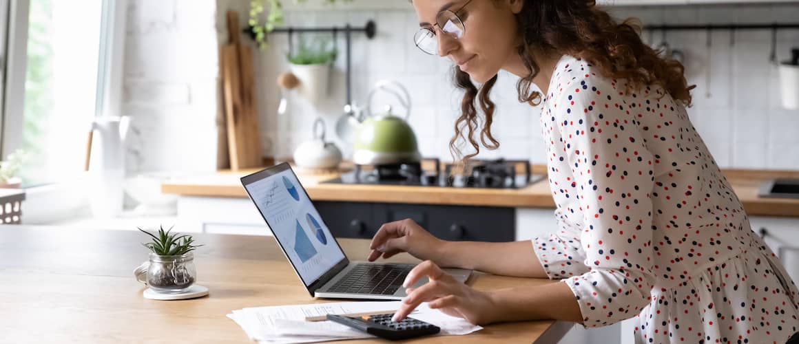 Woman at kitchen counter on laptop calculating finances using online tools and a nearby calculator.