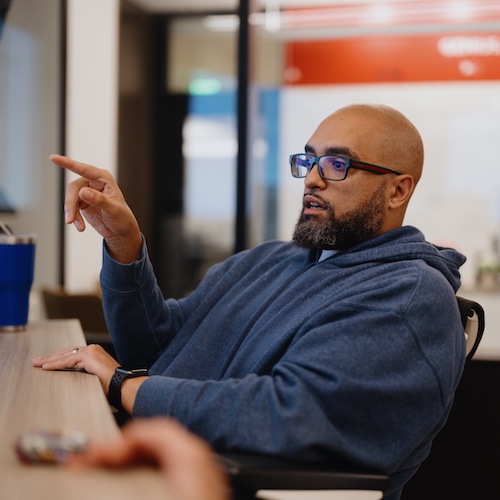 Young man wearing glasses and a blue jacket while sitting at a desk.