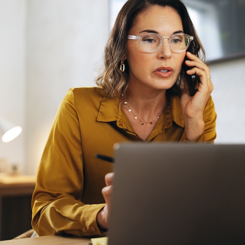 Business woman wearing reading glasses on phone and working on laptop.