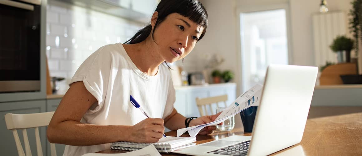 Woman calculating finances at her kitchen table with laptop open and paperwork in her hands.