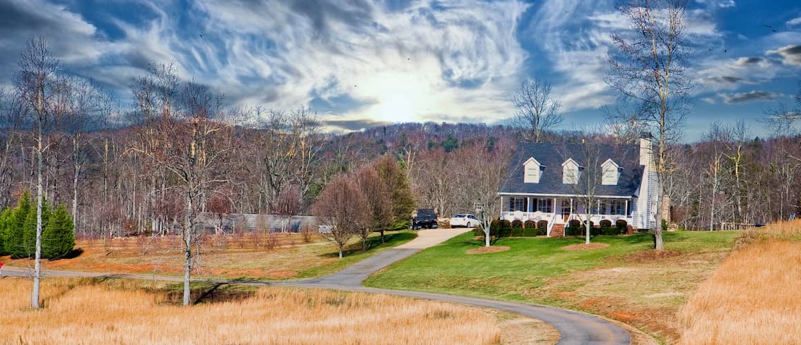 Farmhouse style home on big piece of land surrounded by trees with no foliage in winter.