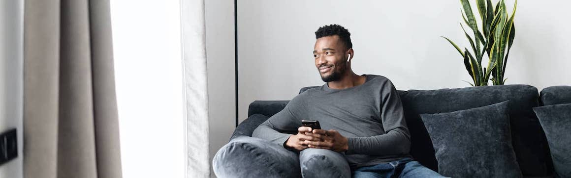 A man seated on a couch with a phone and bluetooth in ear while looking outside the window.