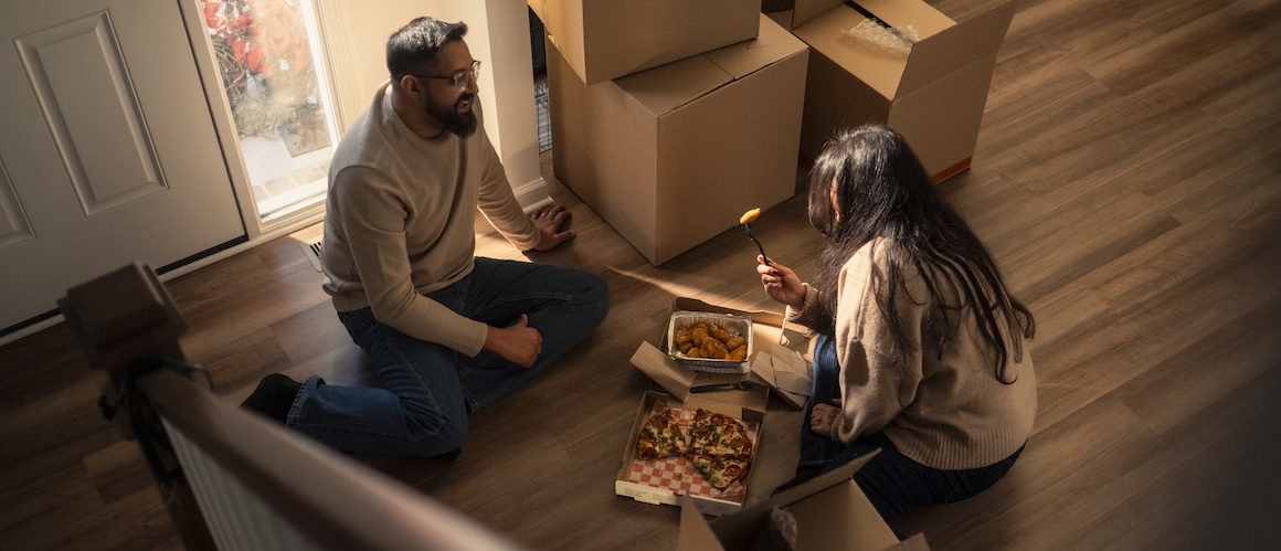 Man and woman sitting on floor of new home surrounding by boxes, eating take out.