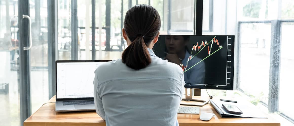 Young business woman looking at multiple computer screens displaying graphs and data in her office.