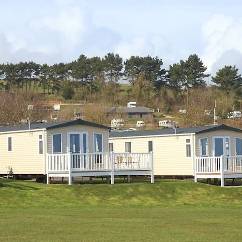 Row of white manufactured homes with expansive green lawn in foreground and trees in the background.