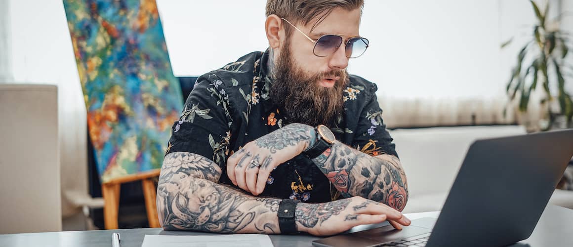 A stylish young man with tattoo sleeves working on a computer.