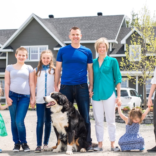Family with five daughters and large dog, possible a military family, in front of grey home.