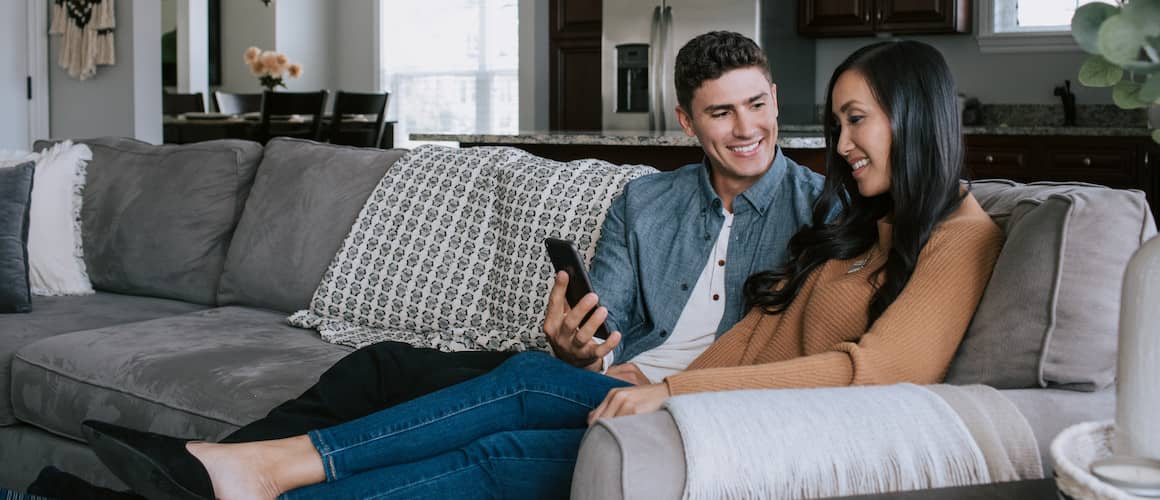 Image of husband sitting on couch, using phone to show personal loan offers to wife sitting next to him.