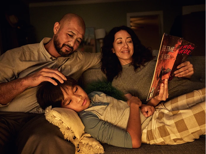 Two parents sitting on a couch with a child resting on a pillow, while the mother reads a book.