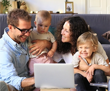 A mother, father and two children laughing and looking at a laptop screen while sitting on the living room floor. 