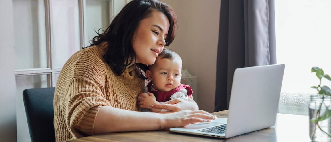 Mother holding child while using laptop at a table.